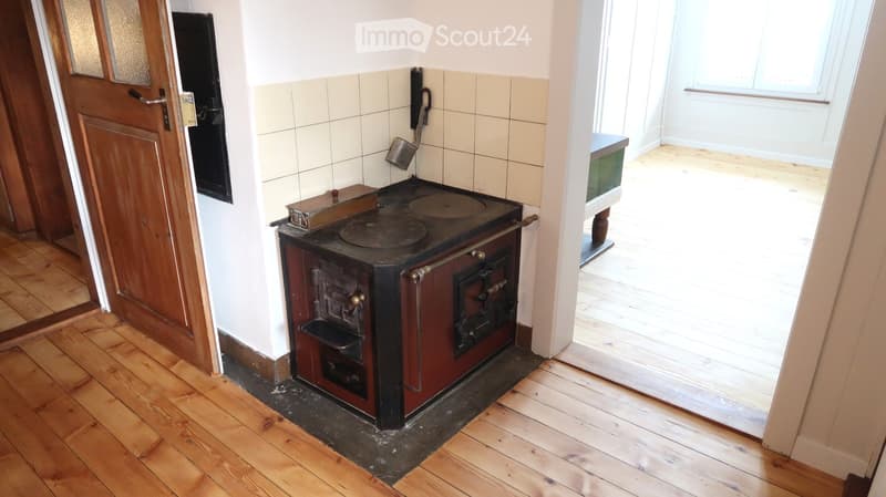 A small kitchen with a wooden floor, a black stove with a pot on it, and a small table with a glass top.