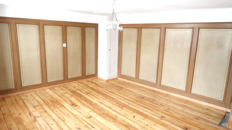 Empty room with wooden panel walls and a polished wooden floor, featuring a chandelier hanging from the ceiling.