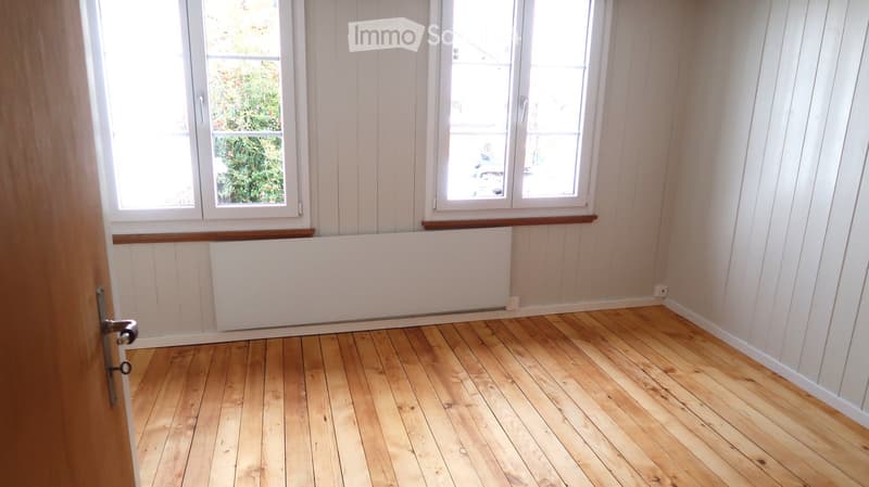 Empty room with wooden flooring, white walls, and large windows allowing natural light. There is a radiator below the windows and a door visible on the left side.