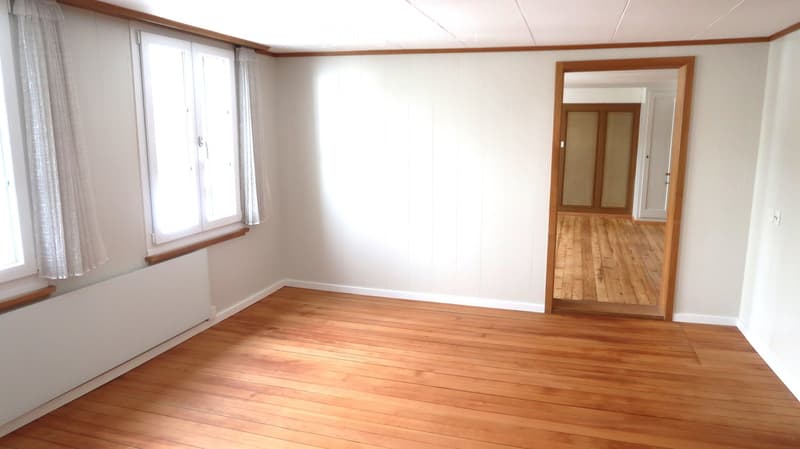 Empty room with wooden flooring, white walls, large window with curtains, and a doorway leading to another room.