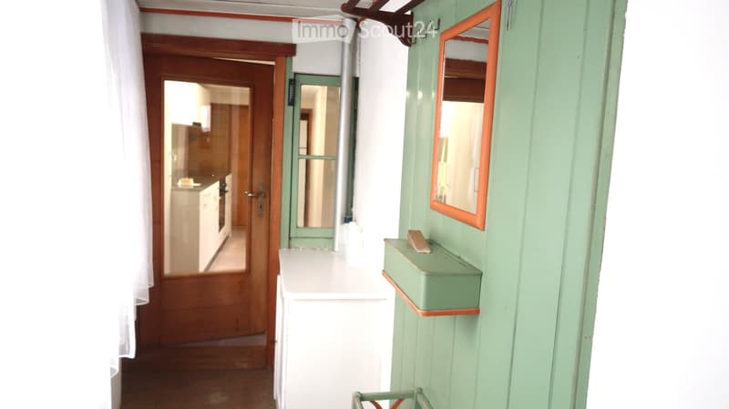 A narrow hallway with light green walls, a small shelf, a mirror, and a door leading to a kitchen area.