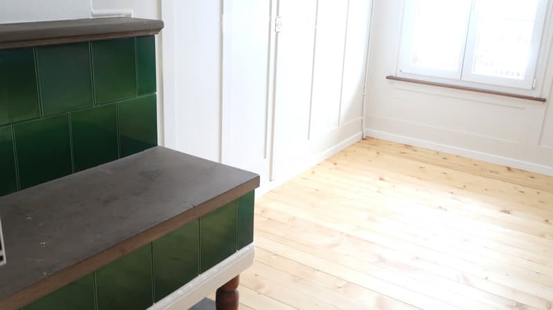 Empty room with wooden flooring, white walls, and a window with a wooden sill. A green cabinet and a dark wooden bench are visible.