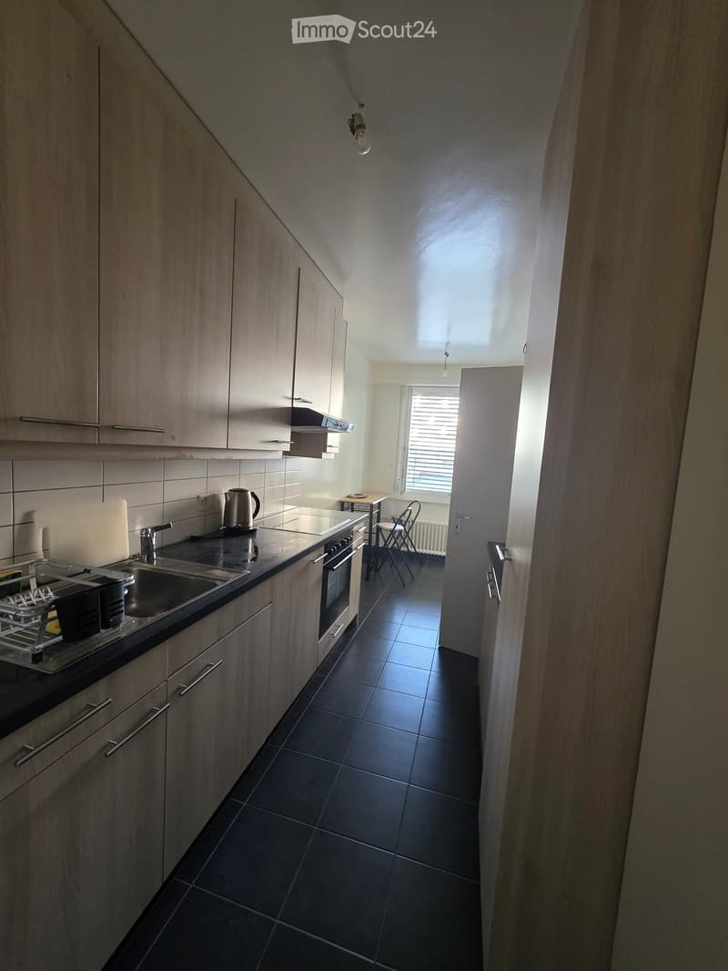 Modern kitchen with light wood cabinets, black tiled floor, stainless steel sink, oven, and small table with chairs in the adjacent area.