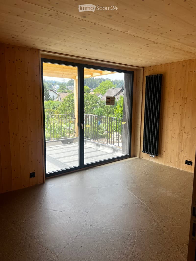 Empty room with wooden walls and ceiling, large sliding glass door leading to a balcony with a view of greenery and houses.