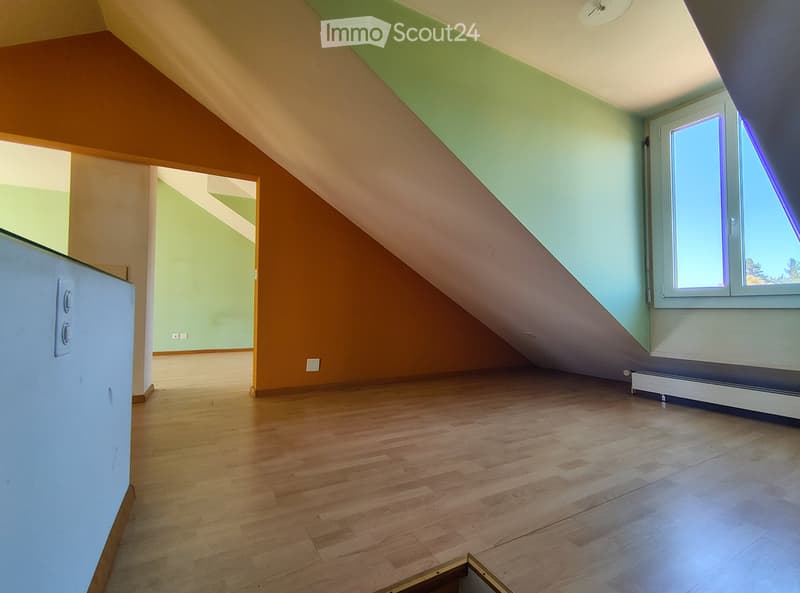 Empty attic room with wooden flooring, slanted ceilings, and large windows allowing natural light. The space is unfurnished and features exposed wooden beams and a doorway leading to another room.
