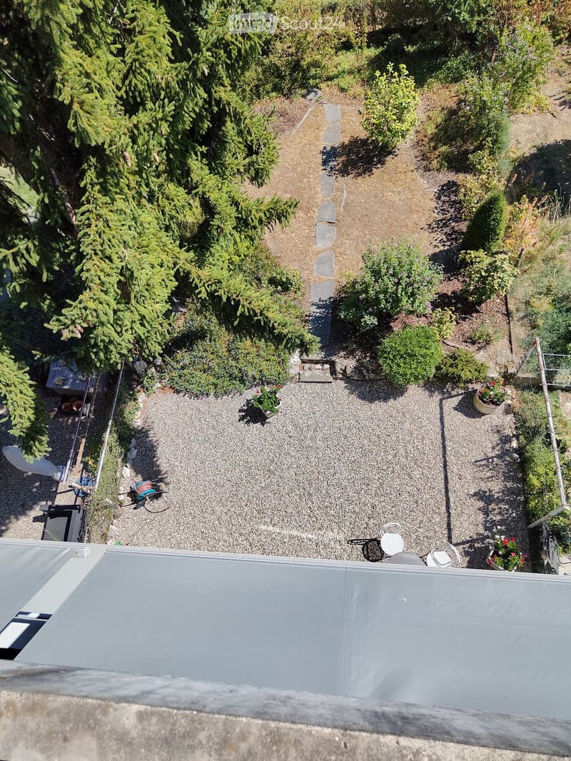 A small, well-maintained garden area with gravel pathways, potted plants, and a few outdoor chairs arranged near a flat roof surface.