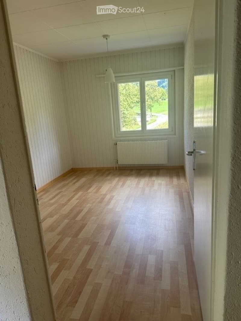 Empty room with wooden flooring, white walls, a window with a view of greenery, and a radiator below the window.