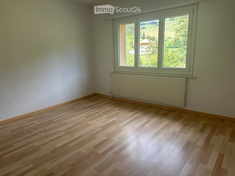 Empty room with wooden flooring, white walls, and a large window with a view of greenery outside. A radiator is visible under the window.