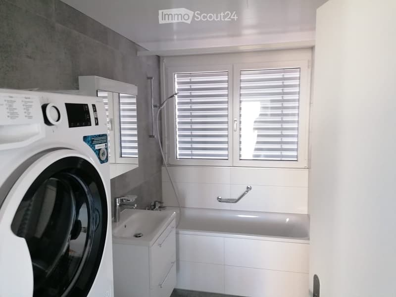 Modern bathroom with a bathtub, sink, faucet, and washing machine. Features white tiles, a window with blinds, and a built-in cabinet.