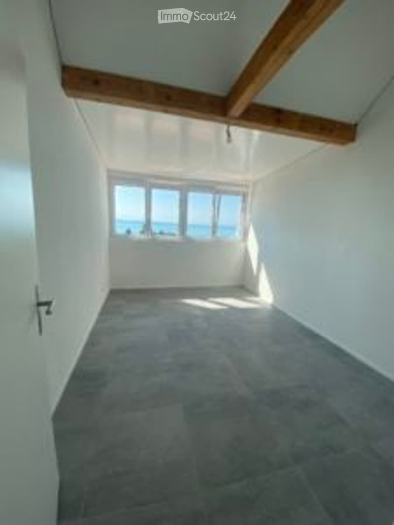 Empty room with white walls, wooden beams, large windows offering a sea view, and a concrete floor.