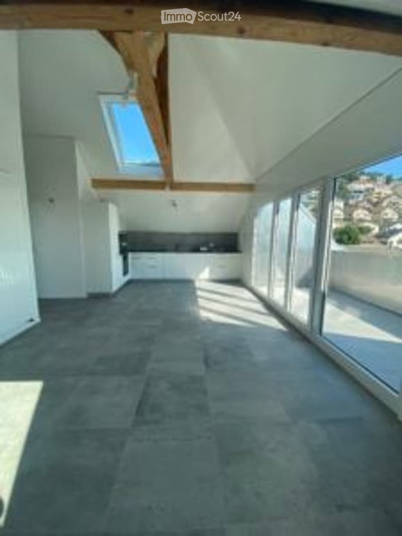 Bright, open attic space with exposed wooden beams, large skylight, and expansive windows offering a view. The floor is tiled, and the walls are painted white.
