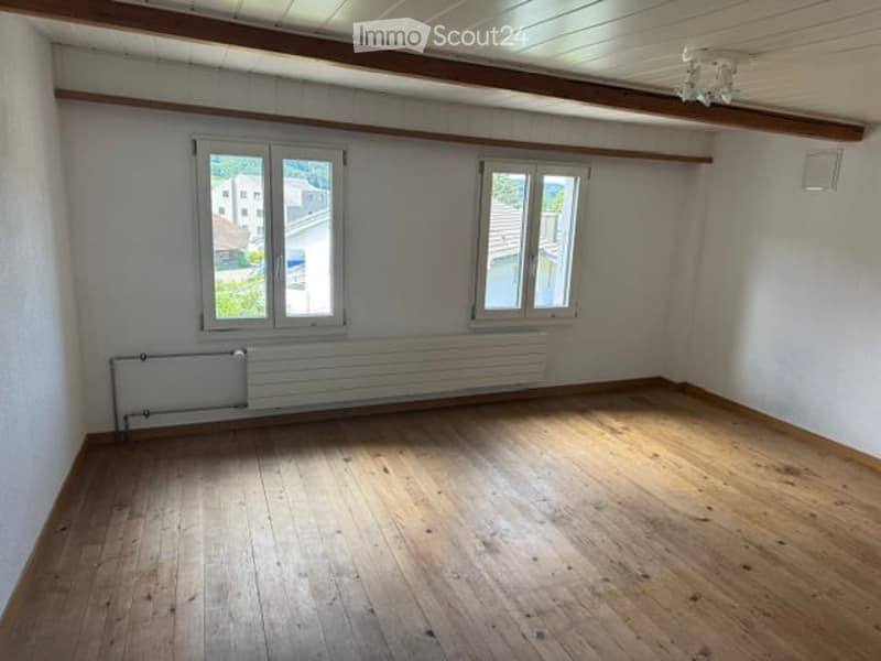 Empty room with wooden flooring, white walls, two windows, radiator, and exposed wooden ceiling beams.