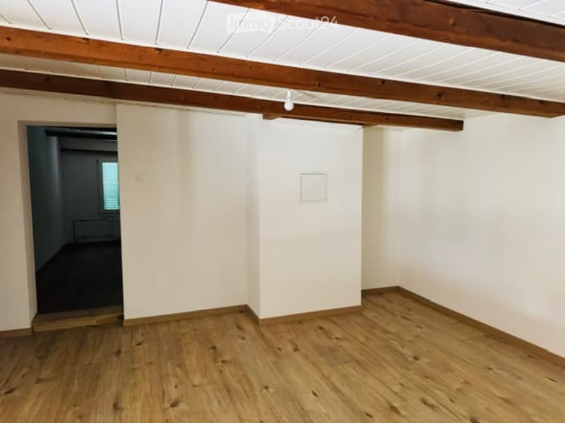 Empty room with wooden flooring, white walls, exposed wooden beams on the ceiling, and a doorway leading to another room.