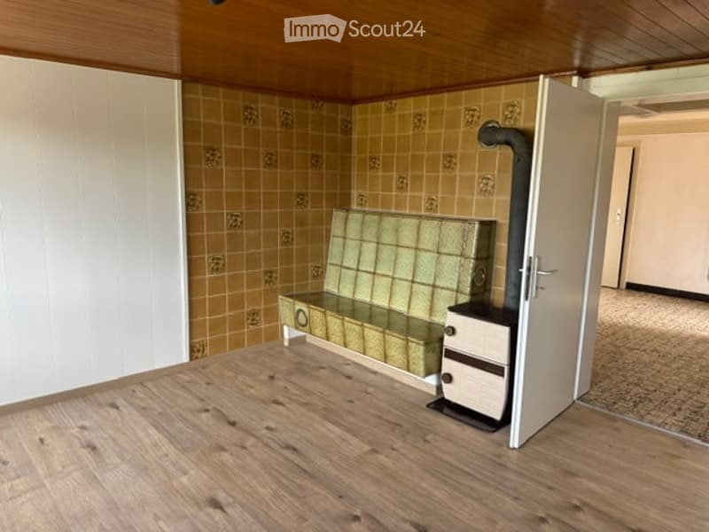 A small, furnished room with a wooden floor, tiled walls, a bench, and a heater. The room has a simple, functional design with a door leading to another area.