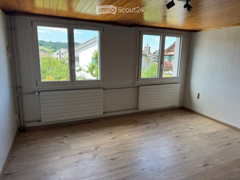 Empty room with wooden flooring, white walls, large windows offering a view of the outdoors, and a radiator along one wall.