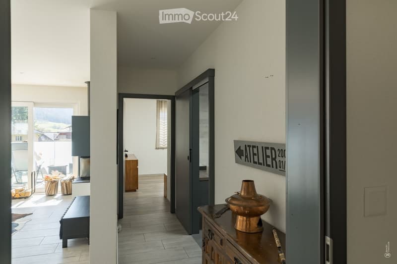 Modern hallway with light-colored walls, wooden flooring, and a decorative copper vase on a wooden console table. A sign reading "ATELIER 201" is mounted on the wall.