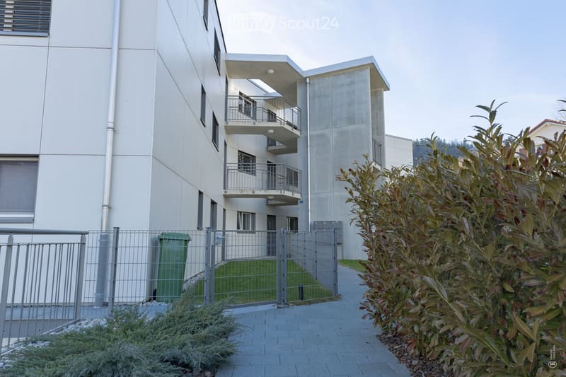 Modern multi-story building with balconies, surrounded by greenery and a gated entrance.