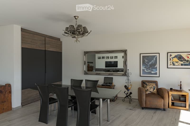 Modern dining room with a glass dining table, black chairs, a laptop on a side table, an armchair, a side table, a large mirror, picture frames, and a decorative chandelier.