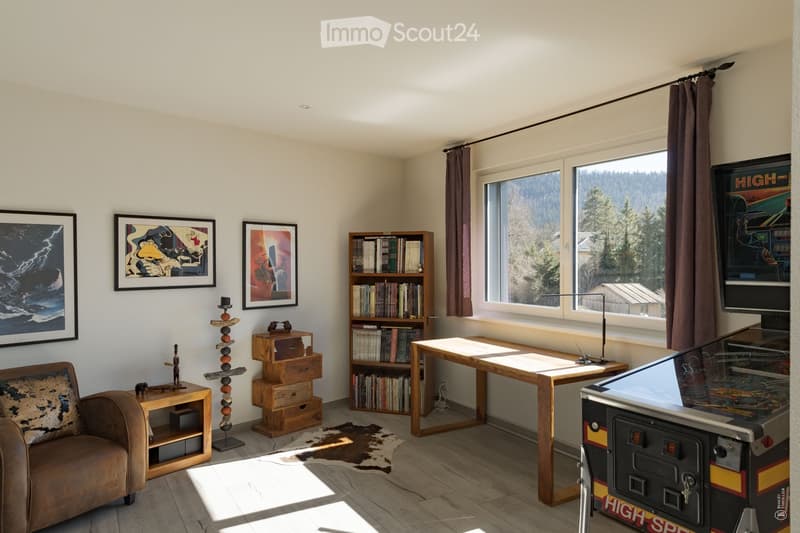 A cozy living room with a brown leather sofa, wooden coffee table, bookshelf filled with books, arcade machine, and decorative items including picture frames and a small wooden stool.