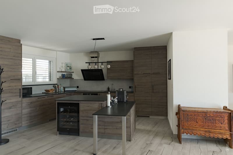 Modern kitchen with wooden cabinets, a central island, wine cooler, microwave, coffee maker, and a decorative chair near the wall.