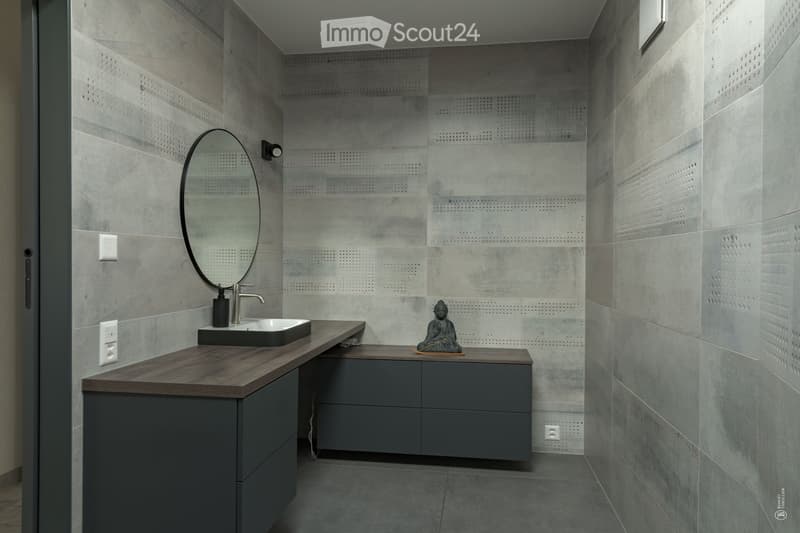 Modern bathroom with grey tiled walls, a dark wooden countertop, a sink, a round mirror, and a small decorative statue on the counter.