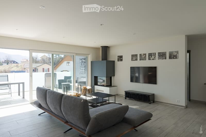 Modern living room with a grey sectional sofa, black coffee table, flat-screen TV, fireplace, and large sliding glass doors leading to a patio with outdoor furniture.