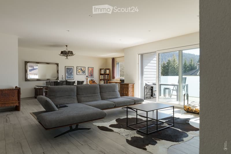 Modern living room with a grey sectional sofa, coffee table, large mirror, bookshelf, dining area with table and chairs, and a balcony with outdoor furniture.