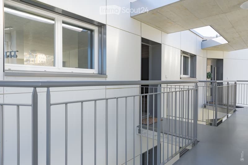 Modern balcony with metal railing, large windows, and a small plant visible in the background.