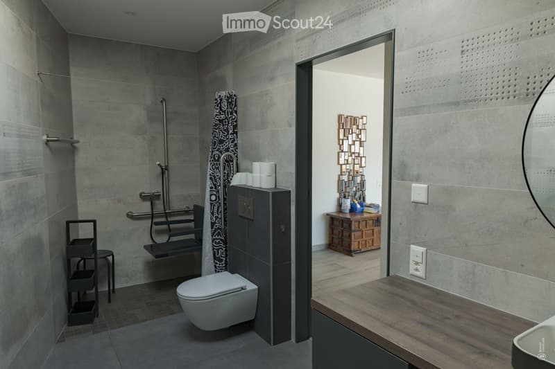 Modern bathroom with grey tiles, a toilet, a shower area with a bench, a large mirror, and a wooden table with a chair in the adjacent space.