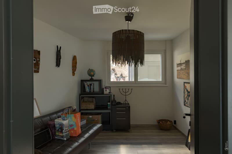 A modern living room with a leather sofa, bookshelf, small table, globe, candle holder, wicker basket, and shopping bags. The room features a large window with a view, a hanging woven chandelier, and several decorative items on the walls.