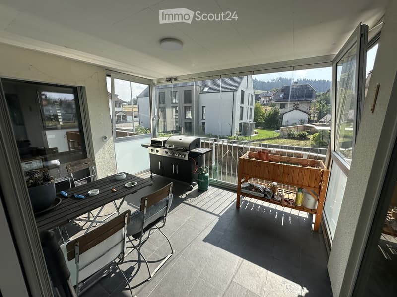 Balcony with a tiled floor, a wooden table with chairs, a barbecue, a wooden planter, and a small bar cart with various items.