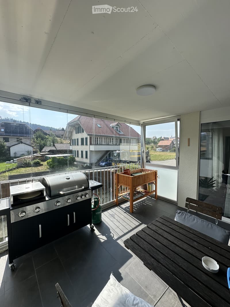Balcony with a modern grill, a small wooden table, and a chair, overlooking a suburban neighborhood with houses and greenery.