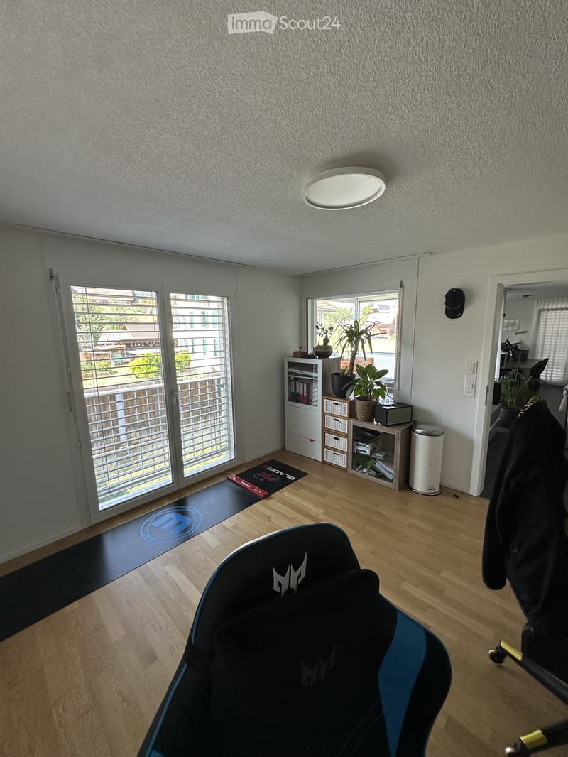 A small, furnished room with a wooden floor, a black gaming chair, a small cabinet with books and a plant, and a trash can. The room has large windows with blinds and a view of the outside.