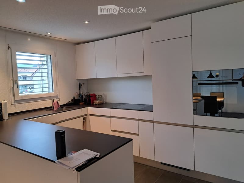Modern kitchen with white cabinets, black countertops, and stainless steel appliances. Features a large island with a sink and a built-in oven. The space is well-lit with recessed lighting and has a window with blinds.