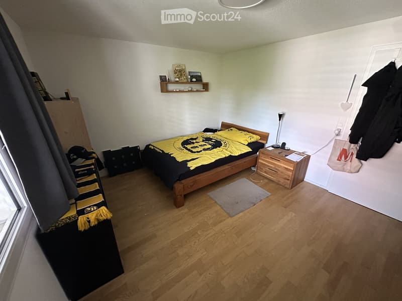Cozy bedroom with wooden flooring, a single bed with yellow and black bedding, a nightstand with a lamp, a small shelf with decorative items, and a rug on the floor.