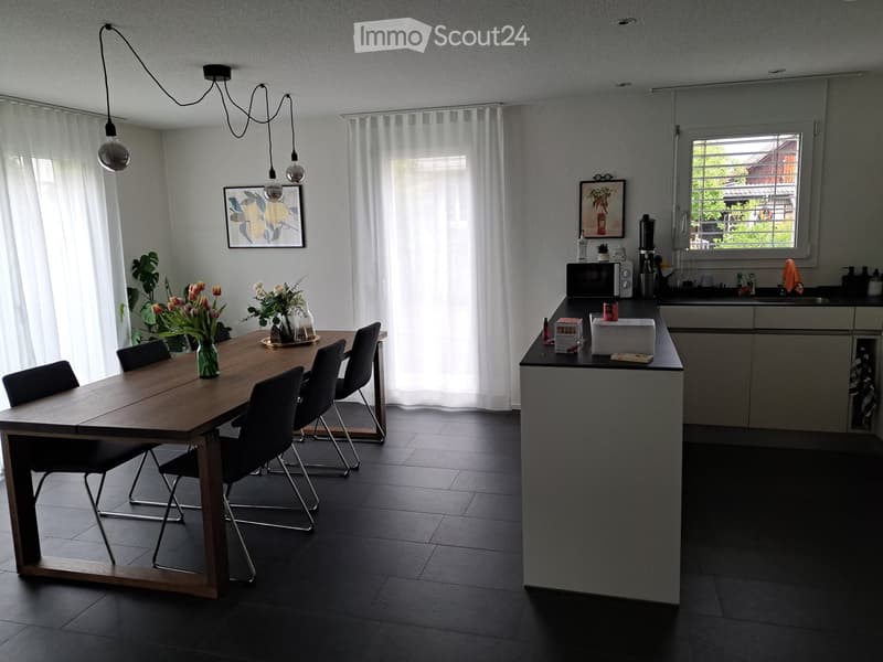 Modern kitchen and dining area with a wooden dining table, four chairs, a vase with flowers, a microwave, and a sink.
