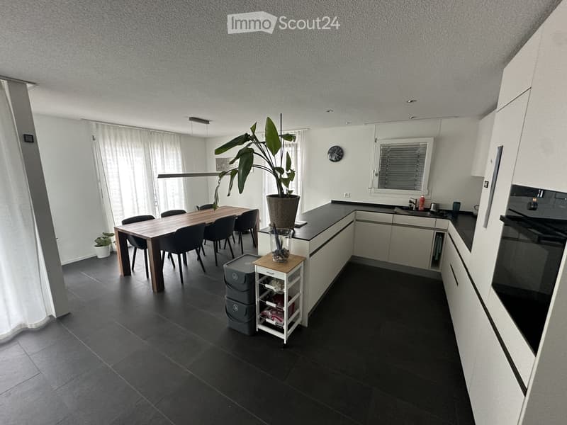 Modern kitchen with dark tiled flooring, white cabinets, a dining table with chairs, a potted plant, and a small wooden stool.