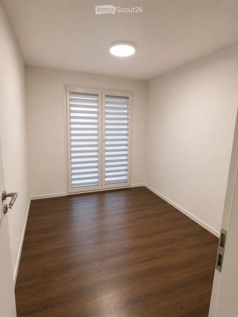 Empty room with wooden flooring, white walls, and a large window with blinds. The room has a single door and a ceiling light fixture.
