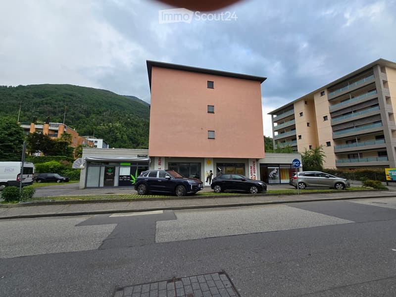 3 story building with a shop on the first floor. Green bushes and mountain in the background.