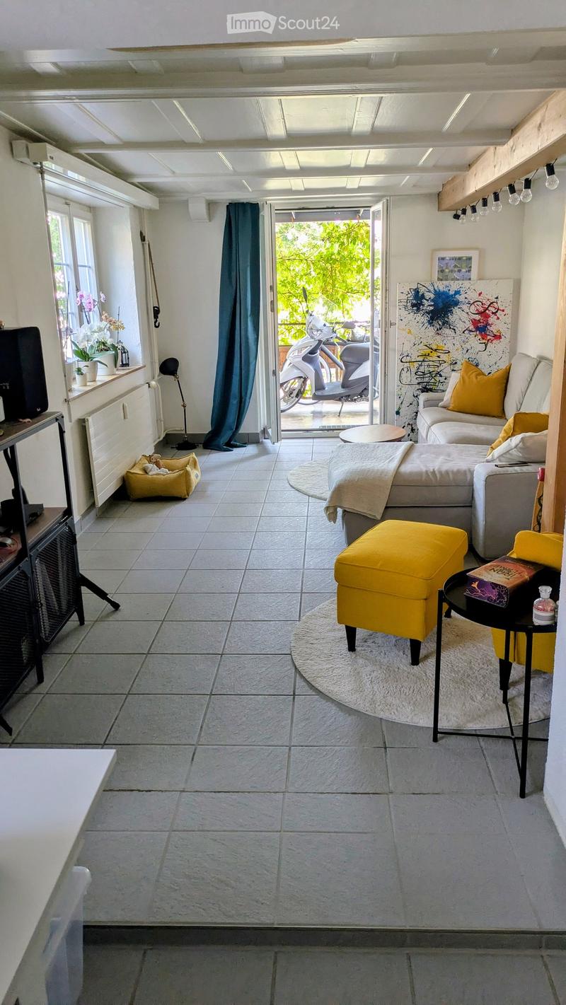 Bright living room with a grey sectional sofa, yellow accent stool, round coffee table, and a black side table with a book. A dog bed is placed near the window, and a large abstract art piece hangs on the wall. The room features a tiled floor and large wi