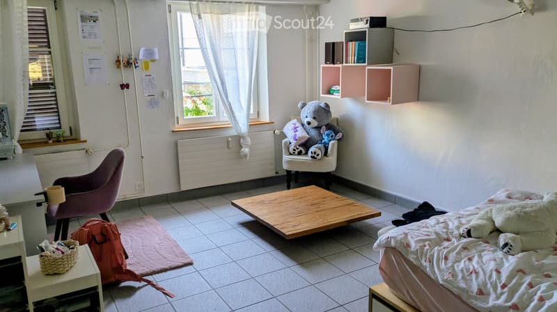 Cozy bedroom with a bed, a wooden table, a purple chair, shelves, and various decorative items including toys and a basket.
