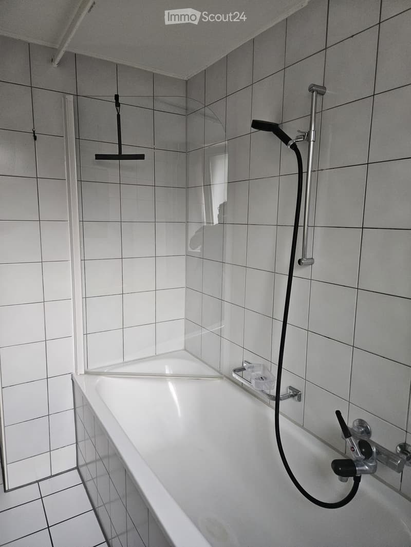 Modern bathroom with a bathtub, showerhead, and a mirror. The walls and floor are tiled in light gray.
