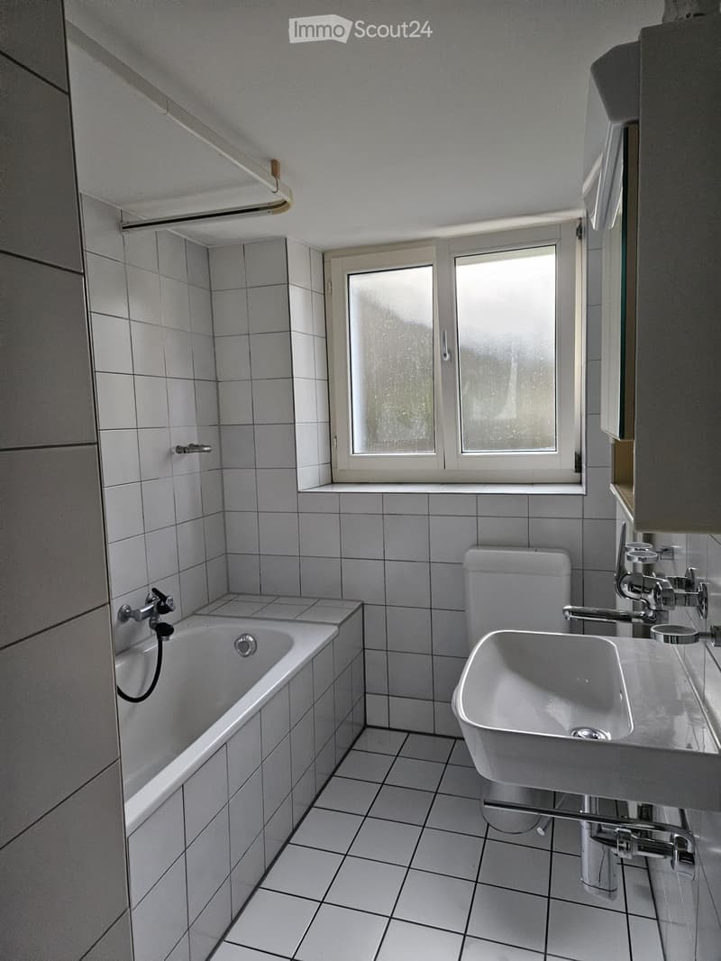 Bathroom with white tiled walls and floor, featuring a bathtub, toilet, and sink. There is a window with frosted glass above the bathtub.