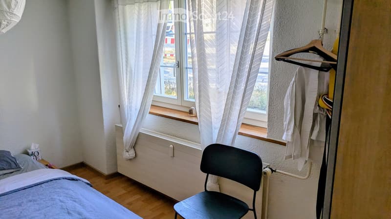 Cozy bedroom with a single bed, a black chair, white curtains, and a radiator. A wooden hanger with a towel is visible on the right side.