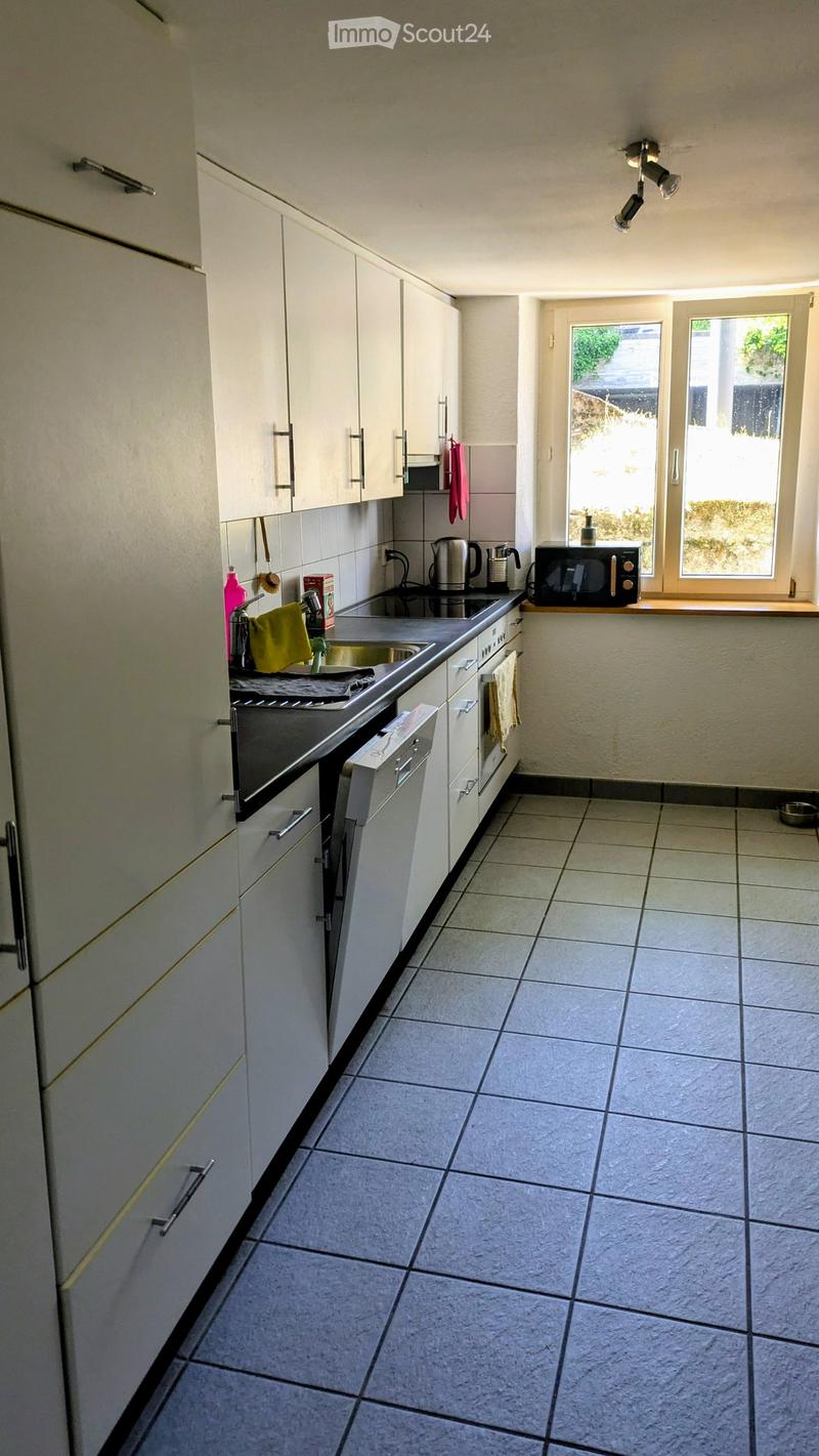 Modern kitchen with white cabinets, tiled floor, and stainless steel appliances including an oven, microwave, and dishwasher. A window provides natural light, and various kitchen items are visible on the counter.