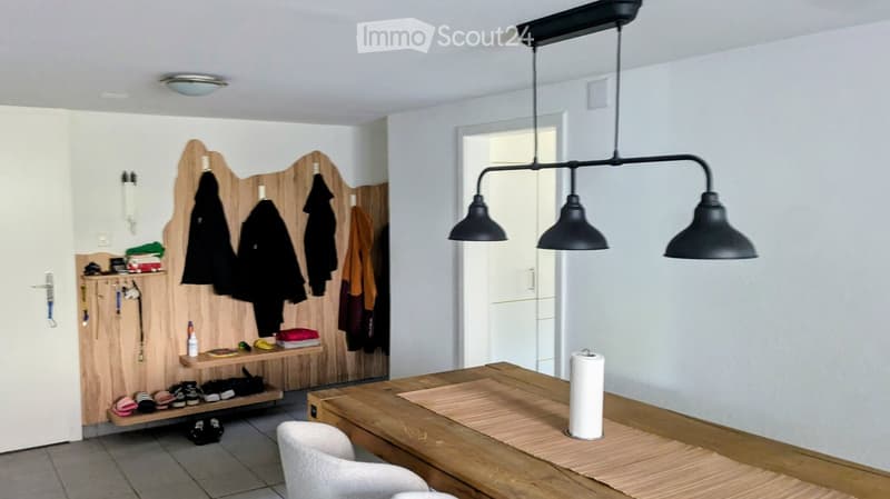 A well-lit hallway or entryway with a wooden coat rack, shelves for shoes and items, a wooden table with a chair, and a modern black pendant light fixture.