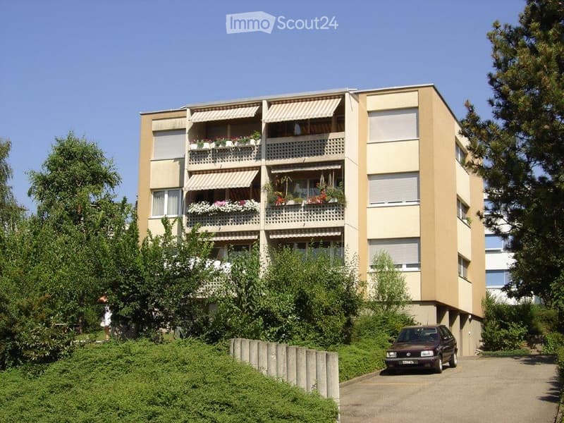 Four-story residential building with balconies, awnings, and flower boxes, surrounded by greenery and a parking area.