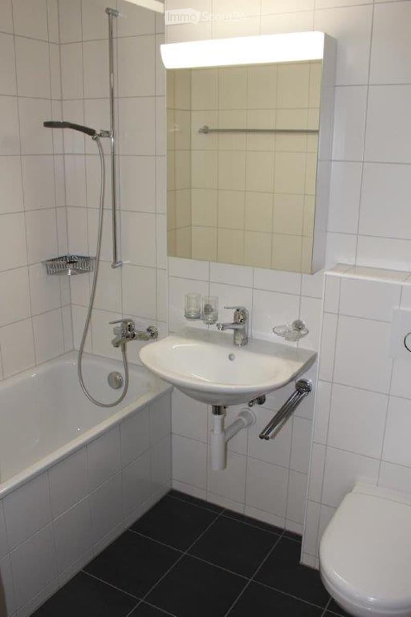 Modern bathroom with white tiled walls, a bathtub, a sink with a mirror above it, and a toilet. The shower hose is mounted on the wall next to the bathtub.
