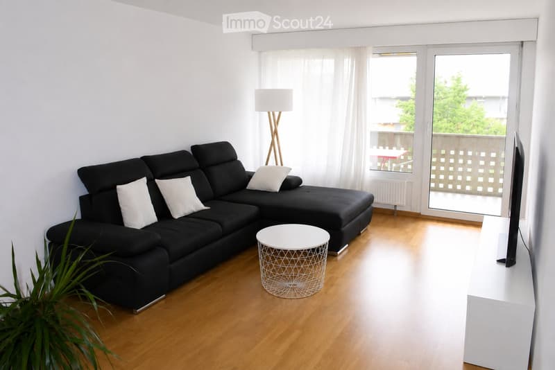 Modern living room with a black sectional sofa, white throw pillows, a white coffee table, a floor lamp, a television, and a small table and chair set visible through the balcony door.