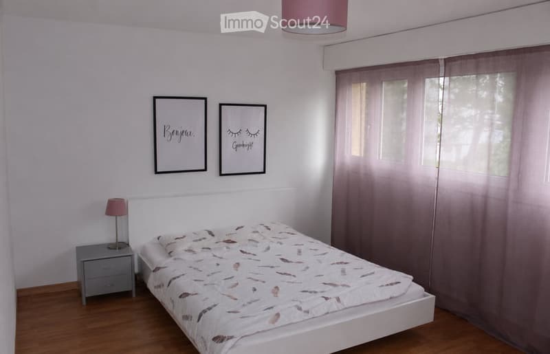 A modern bedroom with a white bed, feather-patterned duvet, nightstand with lamp, and two picture frames on the wall.